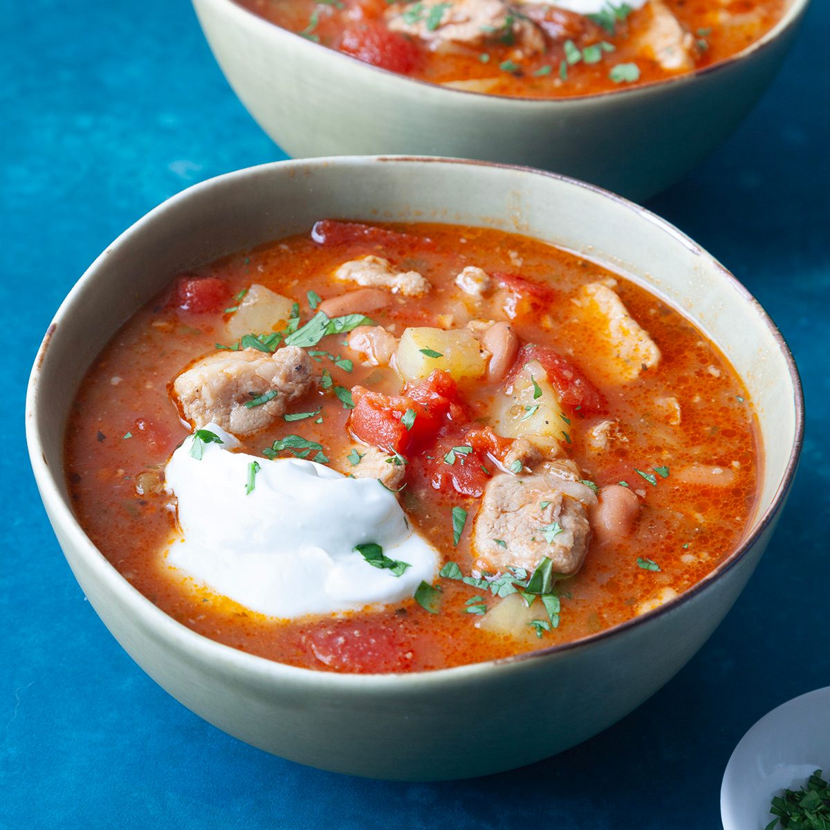 Two single servings of Taste of Home Green Chile Pork Stew in light green bowls on dark turquoise surface, garnished with sour cream and cilantro