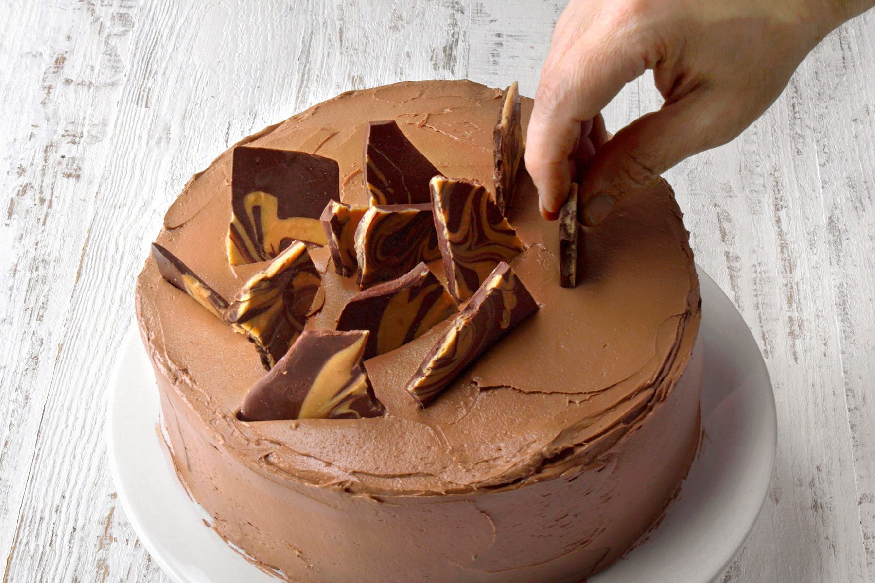 A hand places chocolate shards on top of a frosted chocolate cake. The cake sits on a white plate on a wooden surface. The chocolate shards have a marbled pattern, adding a decorative touch to the smooth frosting.