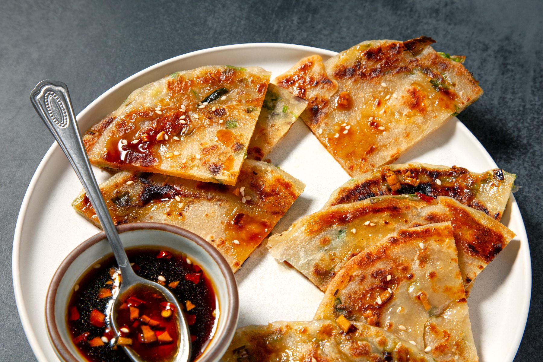 3/4 shot, horizontal orientation, of Chinese scallion pancake pieces on a white plate, accompanied by a small bowl of dipping sauce and a silver spoon, set against a grey background.