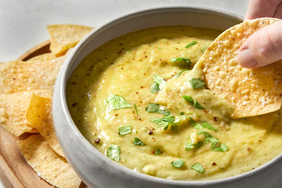 closeup shot of a bowl of Guacamole Salsa, with tortilla chips on the side; a hand is holding a chip, dipping it into the salsa