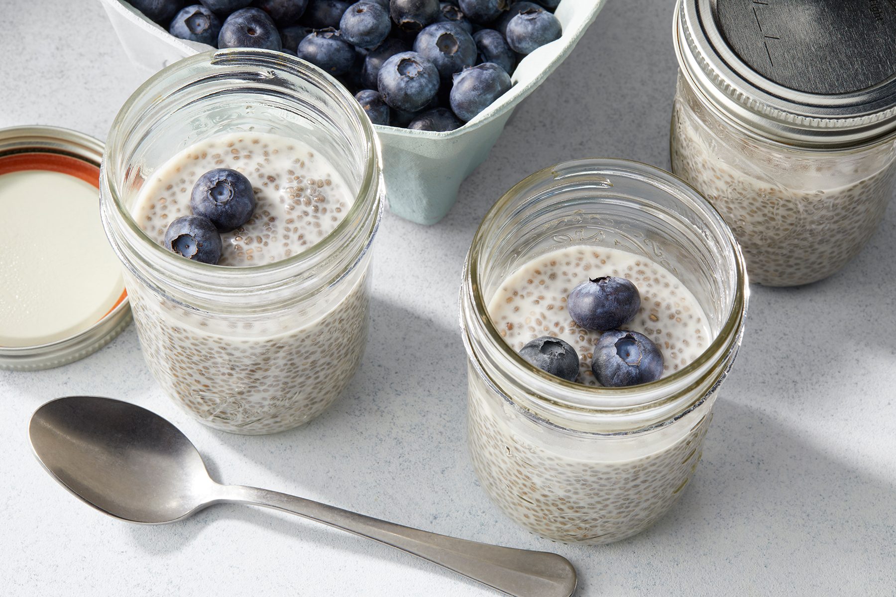 Two glass jars filled with chia pudding and topped with blueberries sit on a white surface. 