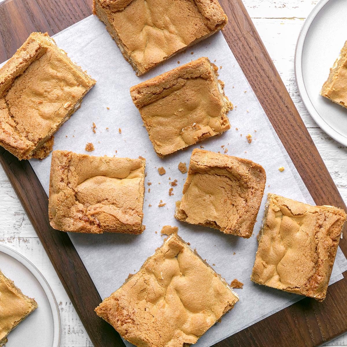 overhead shot of Squares of golden brown blondies are arranged on a parchment lined wooden cutting board with a few pieces on nearby white plates, all set on a white table