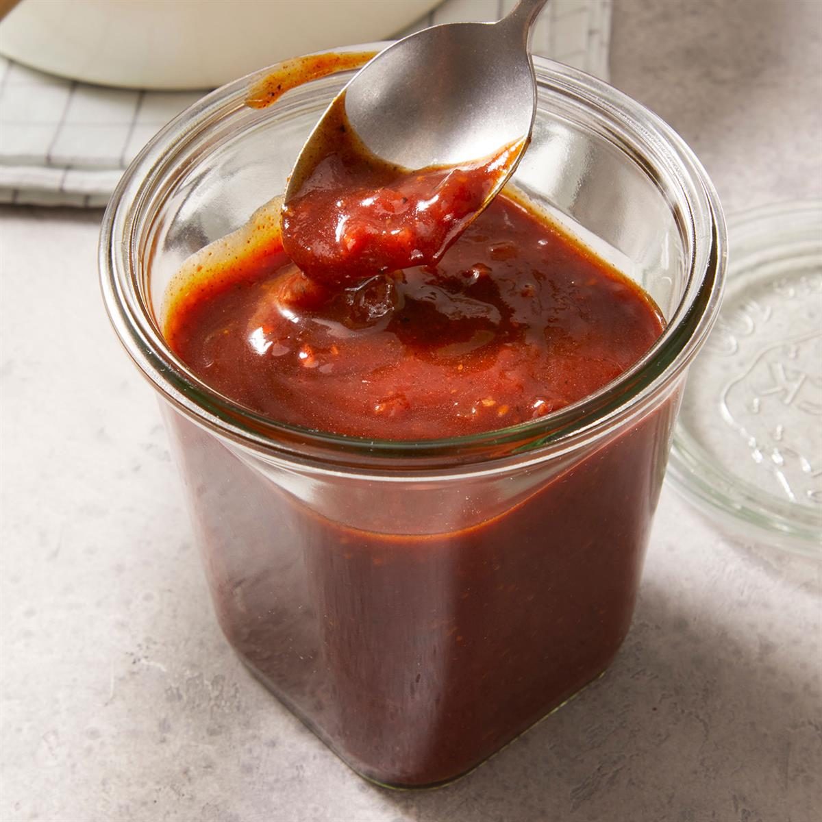 Vertical 3/4 shot of barbeque sauce kept in a glass container with its lid beside it on top of a marble countertop with a check-pattern kitchen napkin kept beside it.
