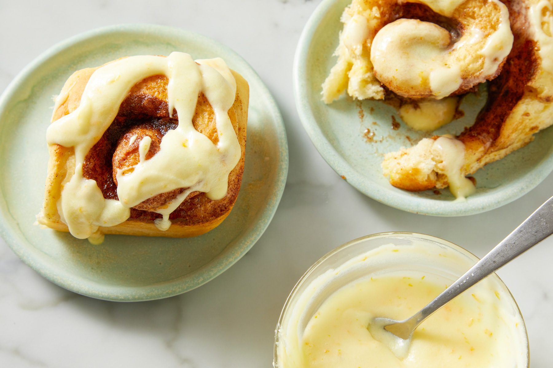 Close-up shot of Best Orange Cinnamon Rolls; served on small plate; Spread prepared frosting over tops; marble surface;