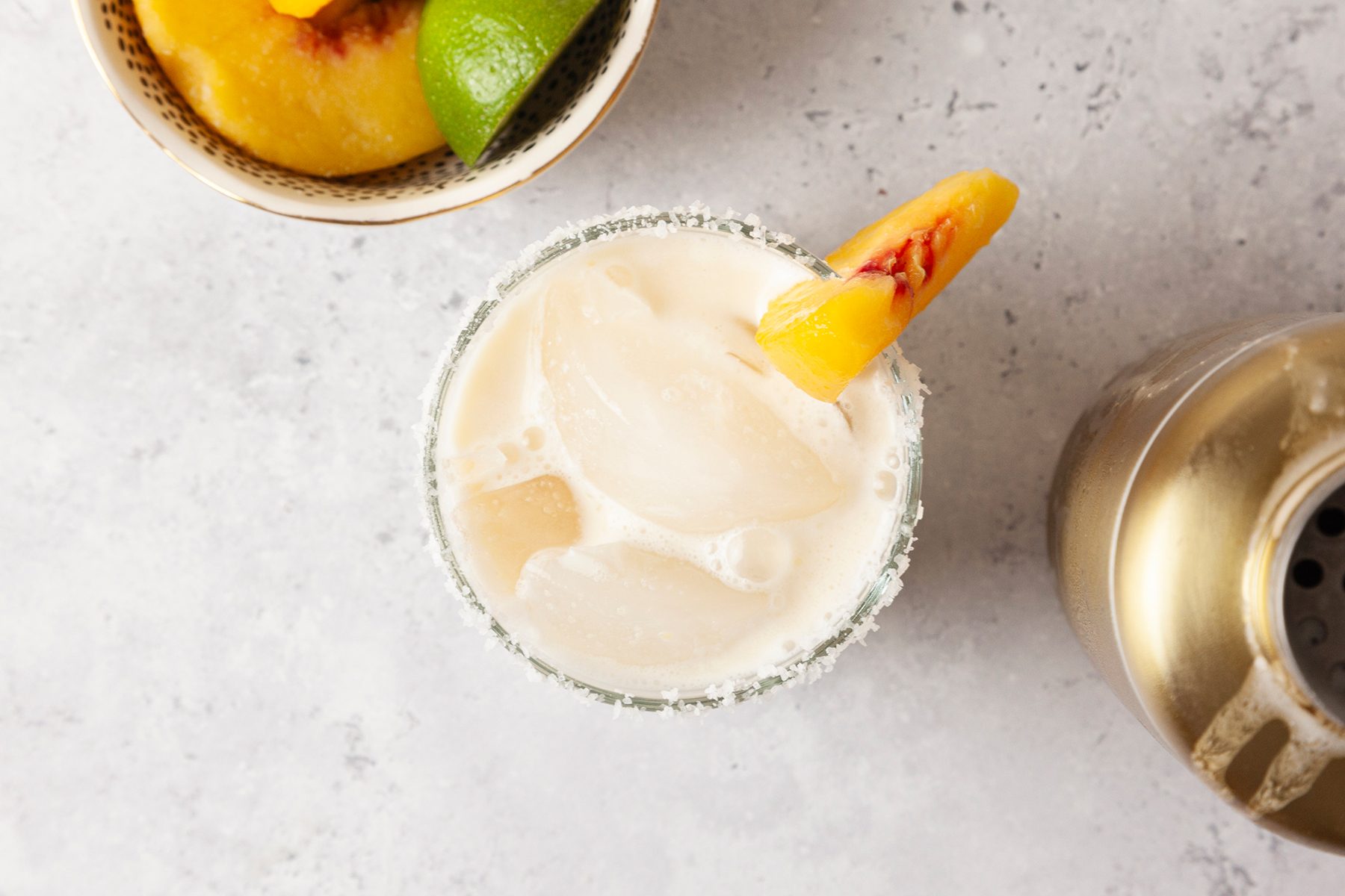 A glass filled with an iced creamy drink, garnished with a peach slice. Next to it are a bowl with peach and lime wedges and a metallic cocktail shaker on a light gray surface.