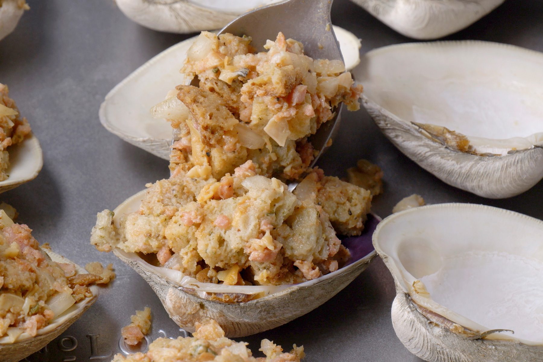 closeup shot of a silver spoon filling an empty clam shell with a light brown and pink mixture, likely a breadcrumb based stuffing containing chopped seafood or meat and vegetables; several other clam shells are visible in the background, some already filled with the stuffing and others still empty; the shells rest on a metallic surface, possibly a baking sheet