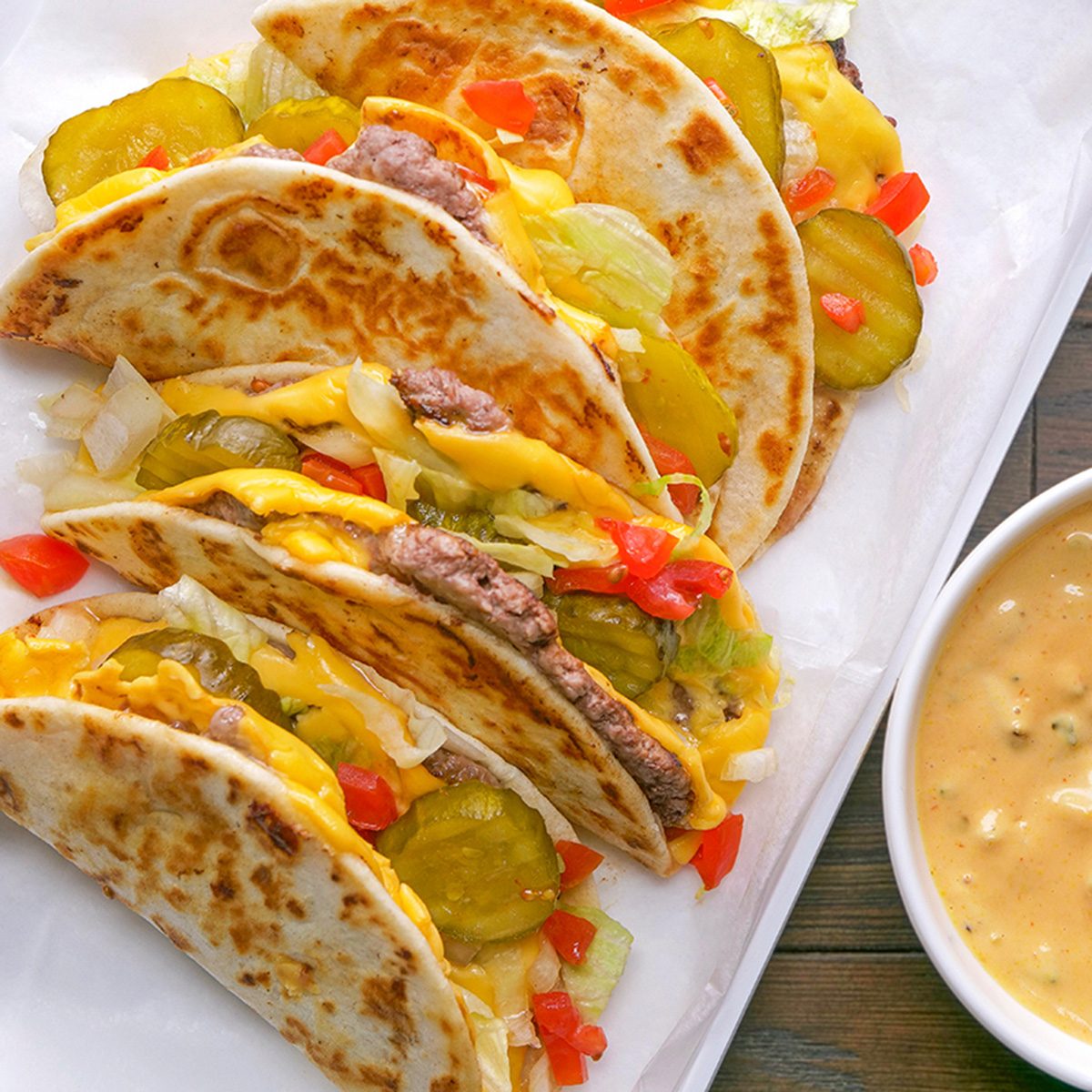 Three cheesy ground beef tacos with pickles, diced tomatoes, and onions are arranged on a white plate next to a bowl of creamy cheese dip on a wooden table.