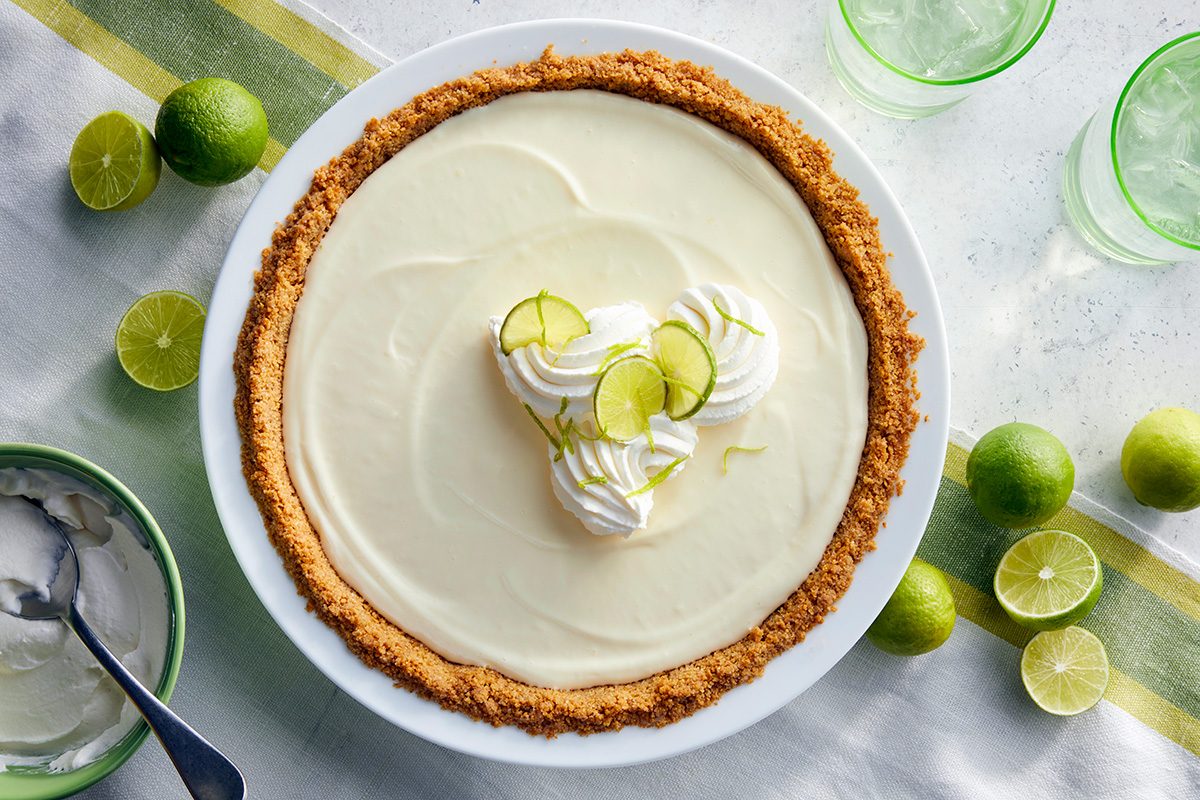 A key lime pie with a graham cracker crust, topped with whipped cream and lime slices, sits on a table surrounded by fresh limes, a bowl of whipped topping, and two green glasses.