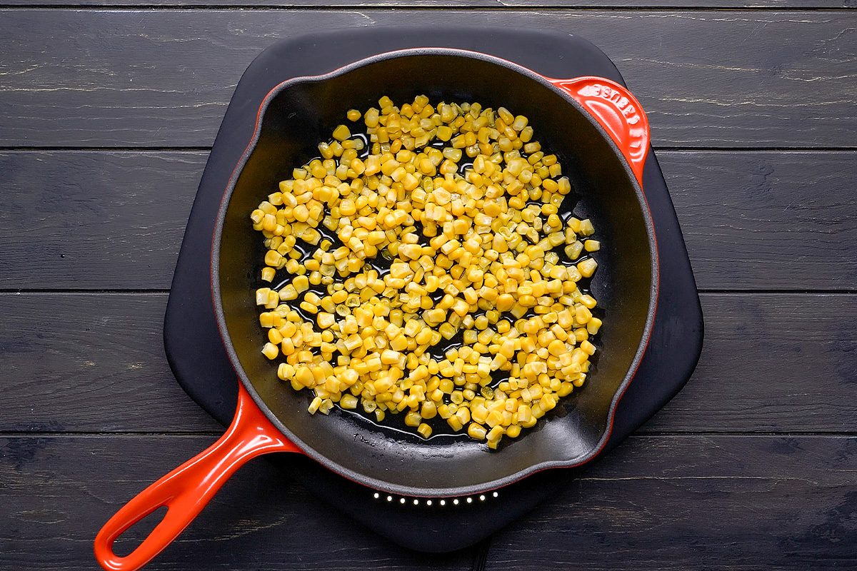 A cast iron skillet with an orange handle sits on a black trivet, filled with cooked yellow corn kernels, on a dark wooden surface.