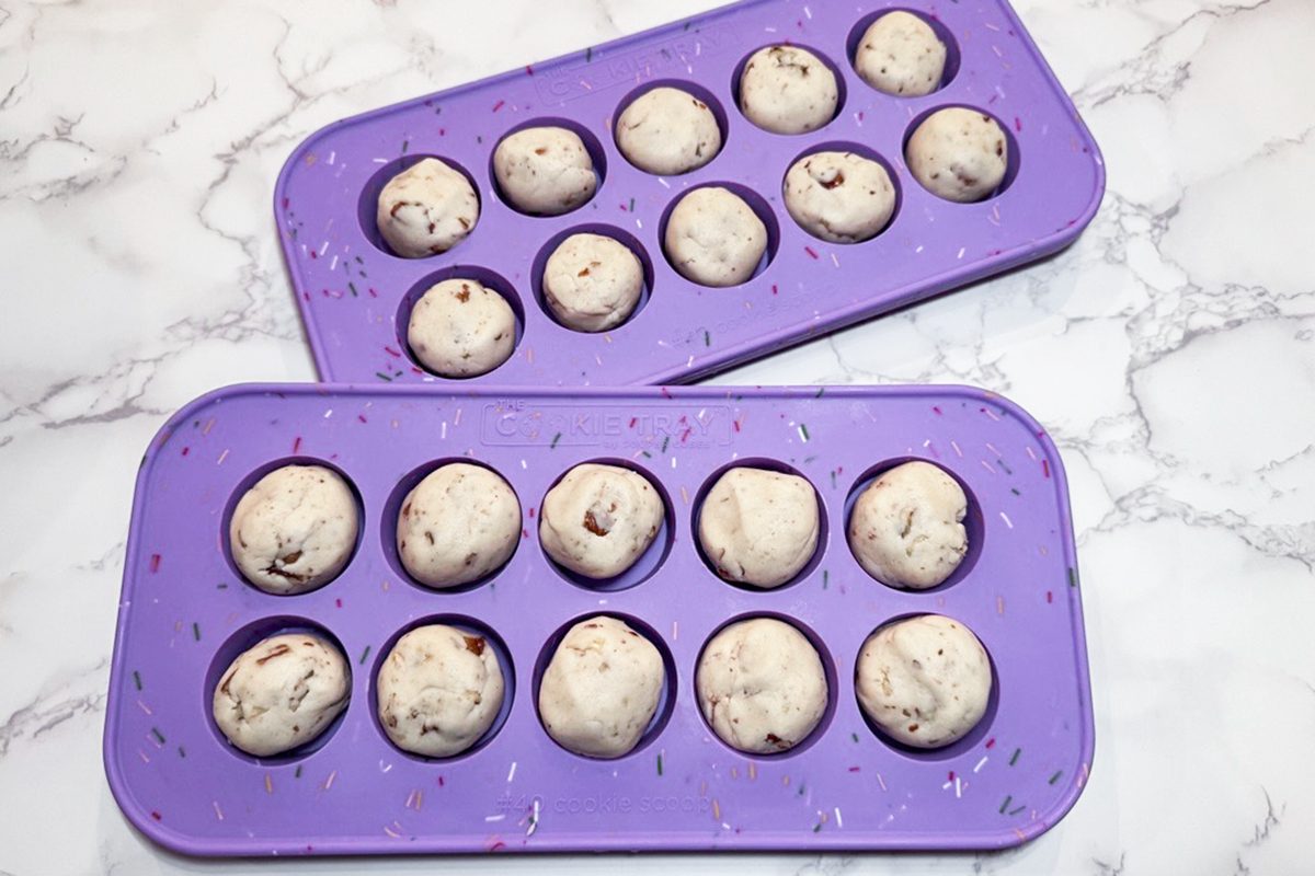 Two purple silicone baking molds, each holding ten unbaked, round cookie dough balls with chocolate chips, are placed on a white marble surface.
