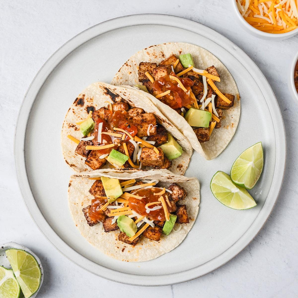 A top shot of a Three soft tacos filled with grilled meat; avocado; shredded cheese; and salsa sit on a plate with lime wedges. Nearby are bowls of shredded cheese; salsa; and a glass garnished with a lime wedge